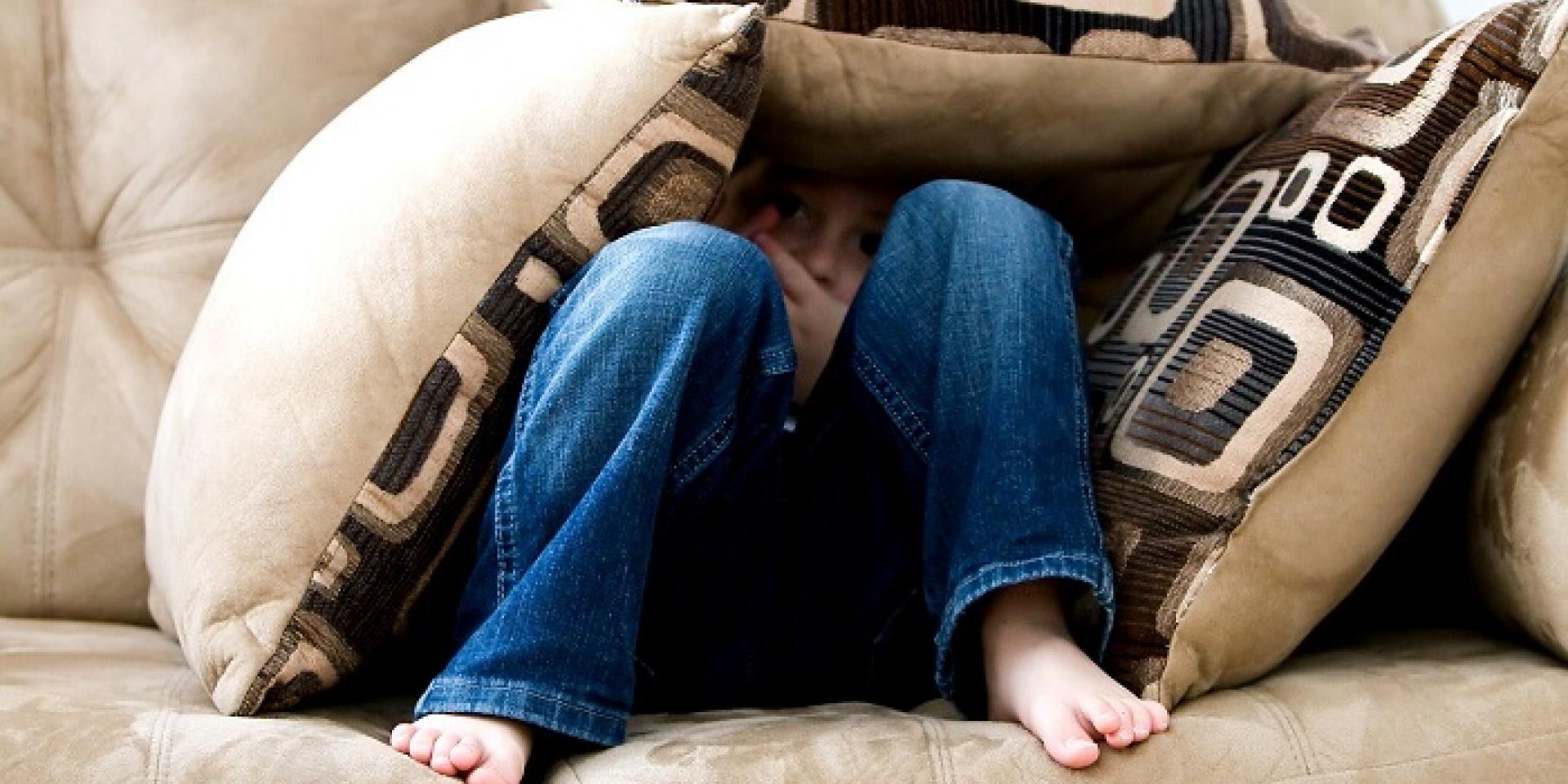little boy sitting on couch with knees up looking ahead mostly covered up with throw cushions