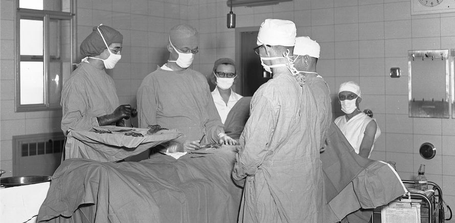 Four masked and gowned people are operating in a historical OR