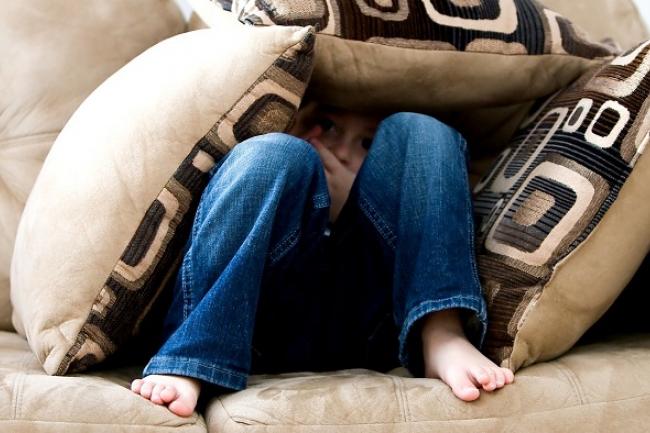 little boy sitting on couch with knees up looking ahead mostly covered up with throw cushions
